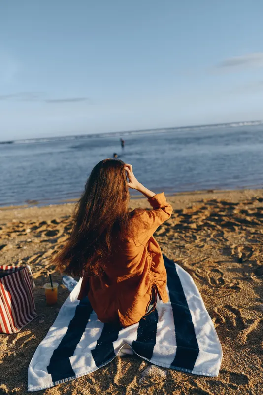 A woman with long hair sits on a striped towel on the sandy beach looking at the calm sea under a clear sky. The scene captures relaxation and peaceful coastal atmosphere.