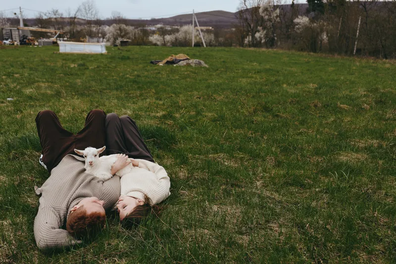 Relaxed outdoor moment with a person lying on grass while a curious goat kid rests on their chest, capturing a simple rural lifestyle, animal companionship, and connection to nature.