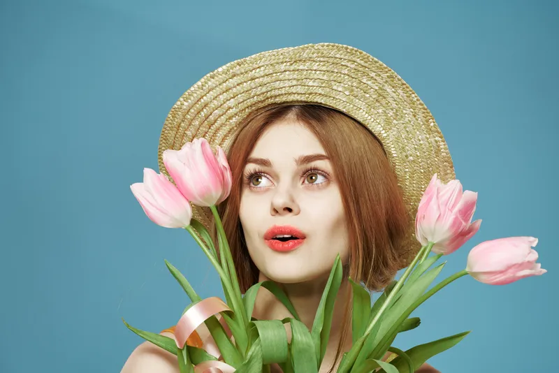 На фотографии высокого качества изображена симпатичная женщина в шляпе с цветами и подарком, что создает атмосферу веселья в честь Международного женского дня