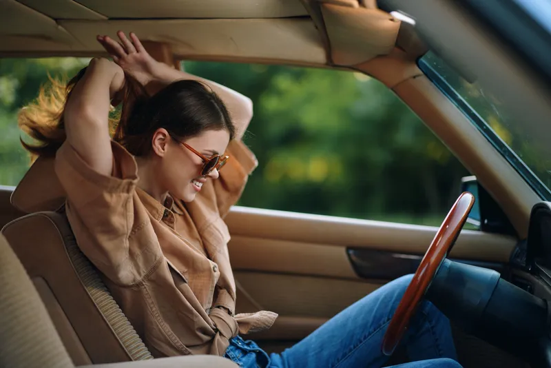 Cheerful young woman with long hair enjoying a drive in a vintage car, wearing sunglasses and a casual shirt, with a lush green background, conveying a sense of freedom and adventure.