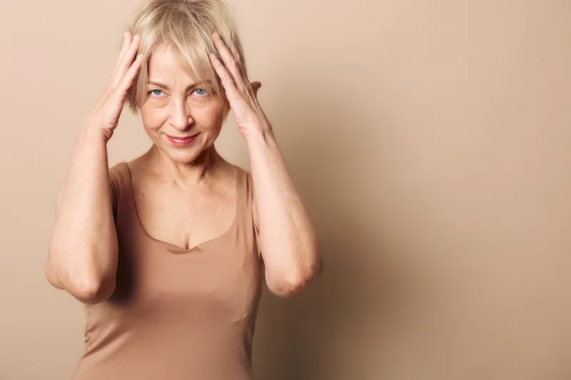 Mature woman with short grey hair touching her head and smiling gently. Neutral beige background and outfit create soft natural mood. Concept of aging, confidence, and wellbeing.