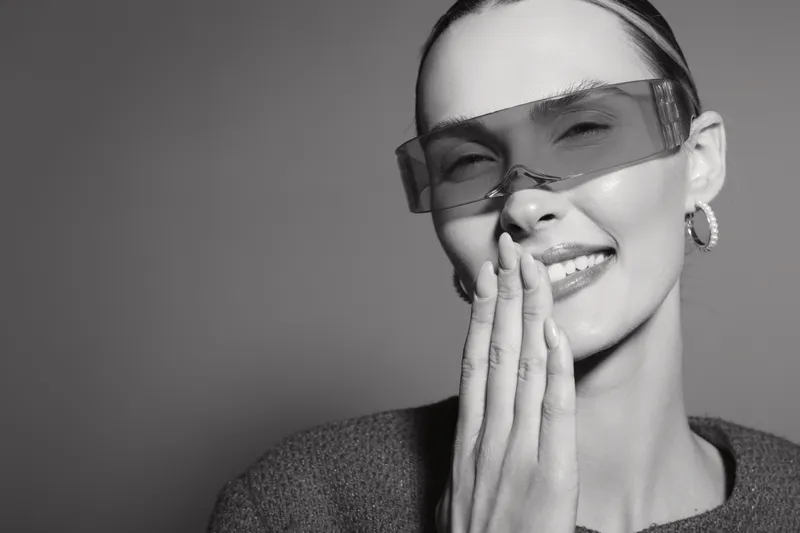 Smiling young Caucasian woman with pale skin wearing futuristic sunglasses and hoop earrings covers mouth with hand. Studio portrait in black and white with soft lighting.