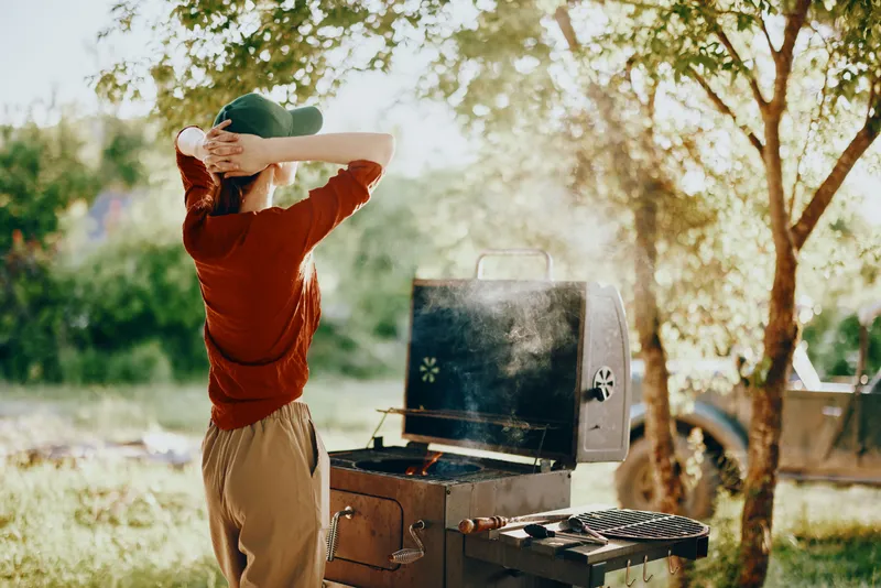 Young woman cooking on a barbecue in a sunny outdoor setting, enjoying a relaxing moment in nature with a joyful expression