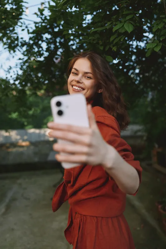 A smiling woman takes a selfie outdoors in a red dress, capturing a casual lifestyle moment in a sunlit park with carefree energy and candid charm