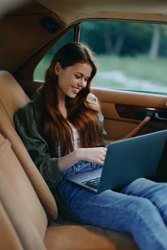Young woman with long hair working on a laptop in a car, smiling cheerfully, dressed casually in jeans and a shirt, capturing a relaxed and modern lifestyle.