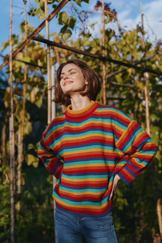 Woman in striped sweater smiling in garden sunlight, young mid-20s with bob haircut and casual outfit. Hands on hips, relaxed joyful portrait in outdoor lifestyle fashion.