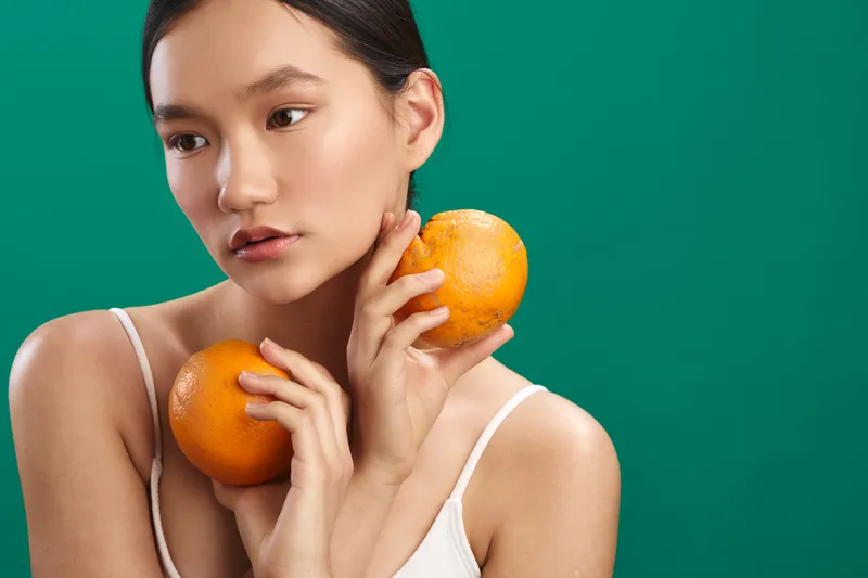 Asian woman holding oranges against a green background, soft lighting, calm expression, highlighting fresh citrus and natural beauty in a simple, modern setting.