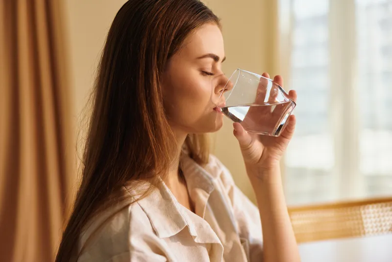 Woman drinking water from glass at home in the morning, side profile portrait showing relaxed hydration routine in cozy interior with natural light, soft calm and healthy lifestyle.