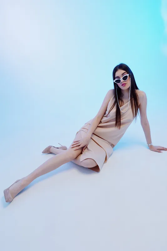 Woman beauty fashion model poses in beige dress and sunglasses on blue background. Studio shot with elegant style and confident mood. Concept for modern fashion and beauty.