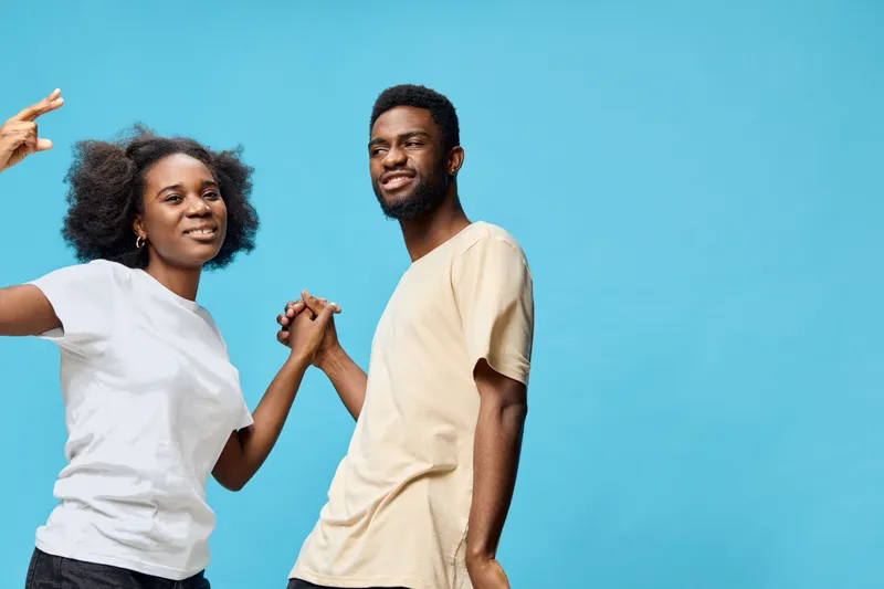 Two young, diverse models dancing joyfully against a vibrant blue background, showcasing happiness and carefree emotions in casual clothing