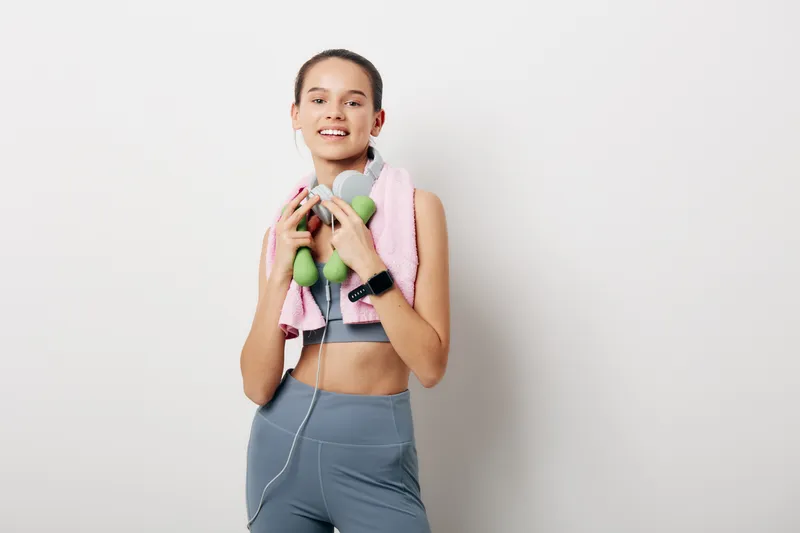 Fitness woman in athletic wear holds small dumbbells and wears headphones with a towel over her shoulders, smiling after a workout against a clean studio background.