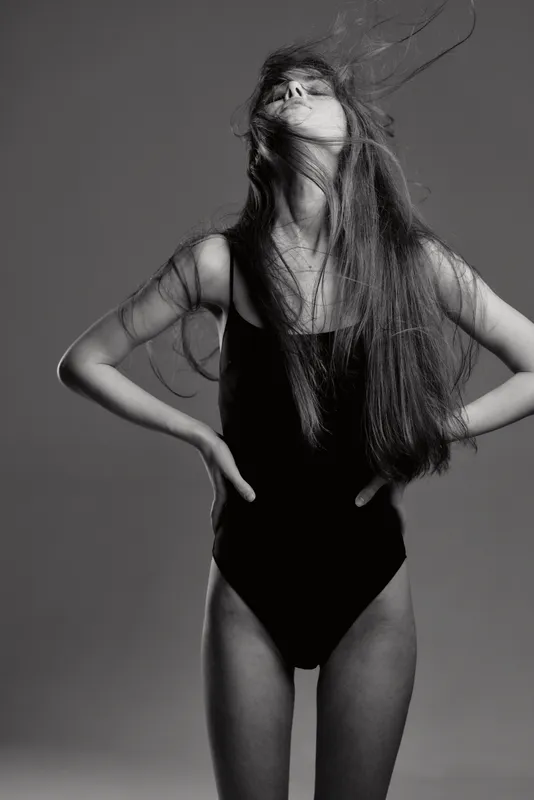 Black and white portrait of a young woman with long hair in a bodysuit, posing with hands on hips and head tilted back in studio.