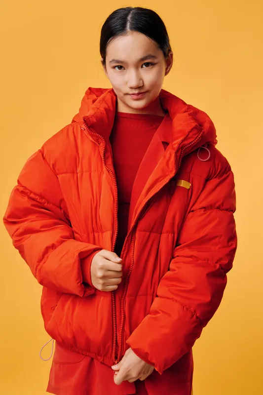 Young woman wearing bright red puffer jacket and matching red outfit standing against orange background. Casual fashion, confident pose with winter outerwear and warm clothing style.