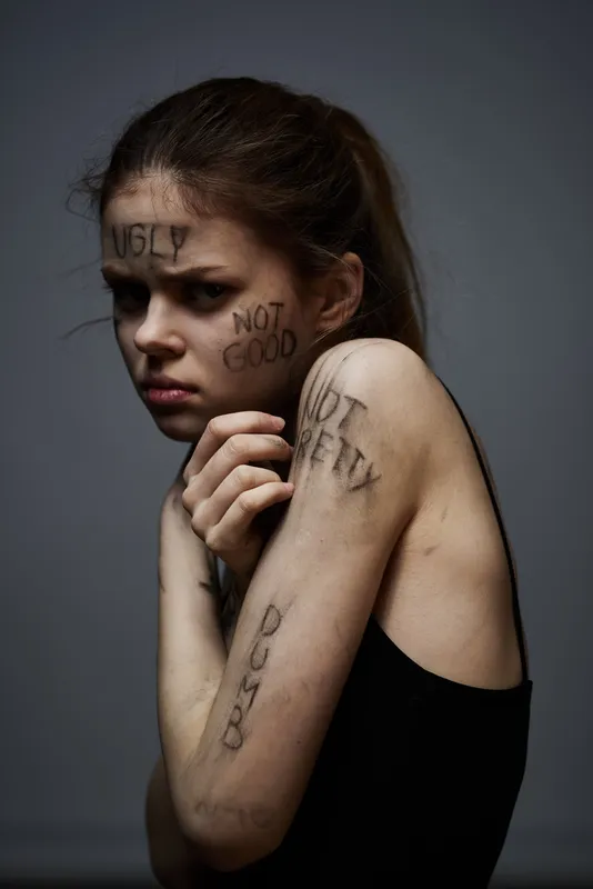 Расстроенная женщина с оскорбительными надписями на теле на сером фоне, касающаяся себя руками, кадр обрезан