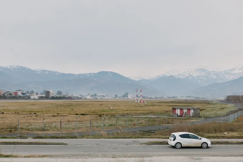 Small white car driving on rural road with dry grass fields and distant snow-capped mountains under overcast sky. Concept of countryside travel, nature, and peaceful landscape scene.