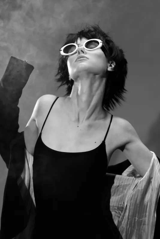 Black and white portrait of a stylish woman wearing sunglasses and a sleeveless dress with expressive pose and short hair.