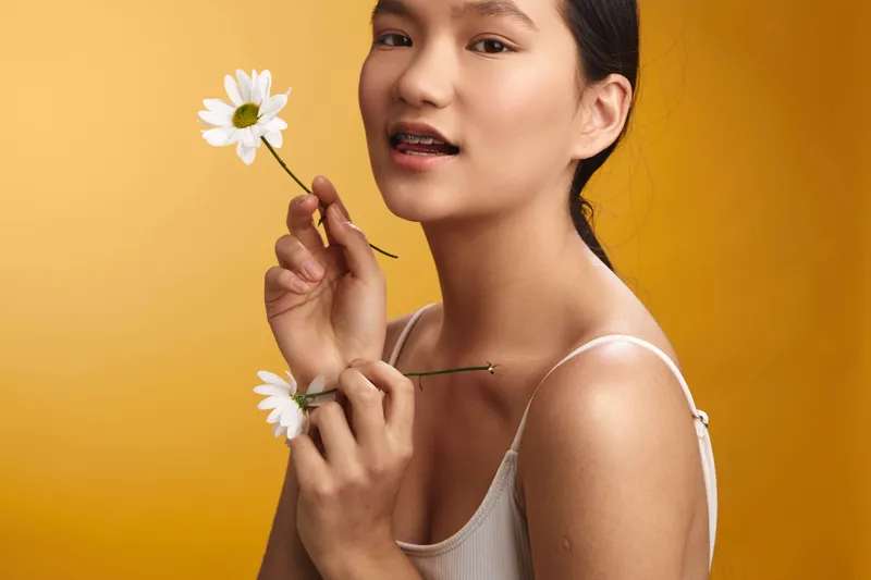 asian woman smiles softly while holding white daisy flowers against a warm yellow backdrop, creating a serene portrait that reflects natural beauty, warmth, and quiet charm.