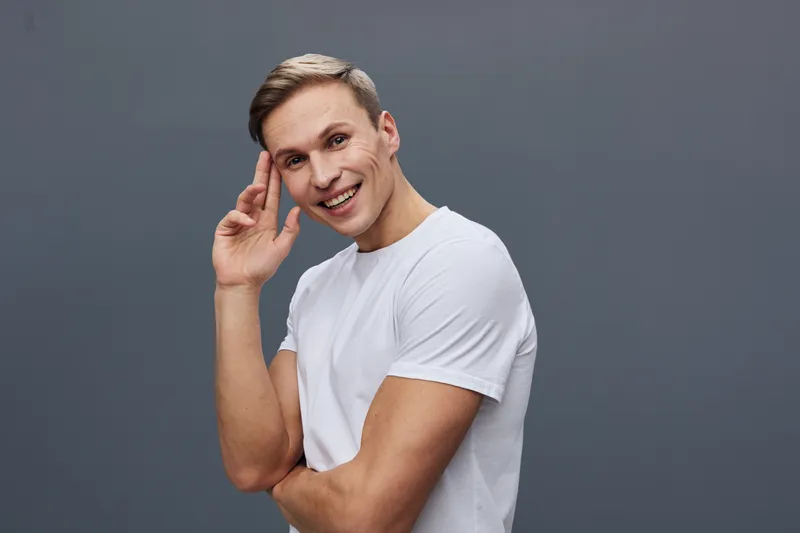 Smiling young man with short blond hair wearing casual white t-shirt, looking confidently at camera, hand near head, cheerful expression isolated on plain gray background, studio portrait, people life