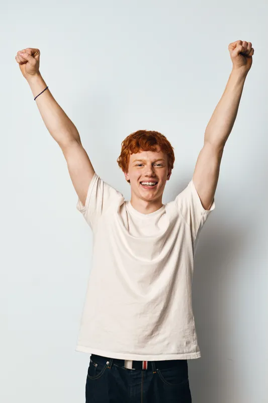 Young man with red hair smiling and raising both hands in victory gesture against white background. Casual clothing, happy expression and energetic body language of male person.