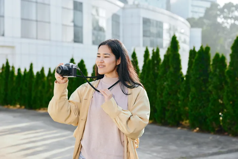 Smiling woman holding a camera outdoors in a casual jacket and sweater, daylight urban setting, candid photography vibe for lifestyle, travel and creative content.