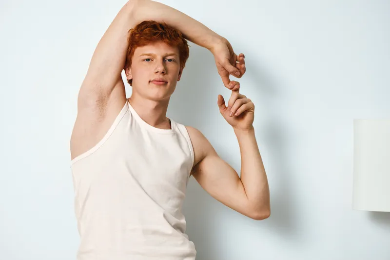Young man with red hair stretching arm while standing against white wall, wearing white tank top in casual indoor setting with lamp in background, portrait with natural light.