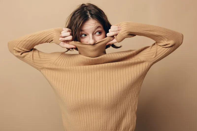 Young woman in beige sweater pulls collar over her mouth, looking sideways with playful expression. Studio portrait on neutral background with casual fashion and cozy mood concept.
