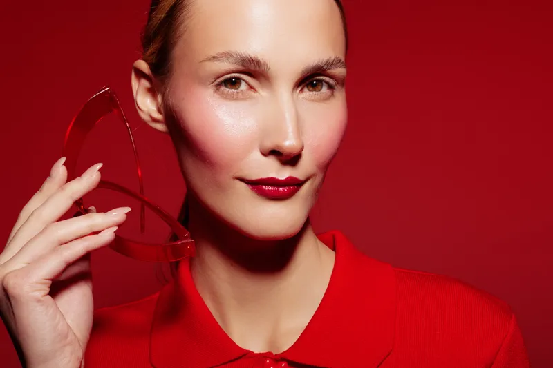 Portrait of confident woman with red lipstick and blush holding transparent glasses against red background. Concept of fashion, beauty, makeup, and style with vibrant color contrast.