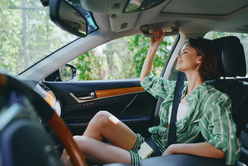 Woman sits in a car seat adjusting the sun visor and mirror, wearing a green striped shirt, soft daylight, calm and candid travel moment.