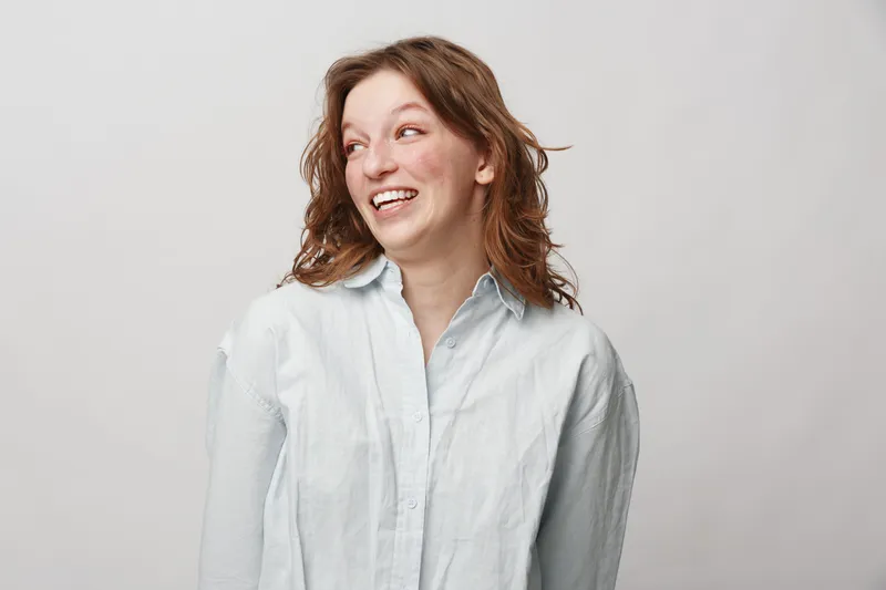 Smiling woman with curly hair wearing white shirt looks away laughing. Studio portrait on light background. Concept of happiness, joy, and natural beauty.