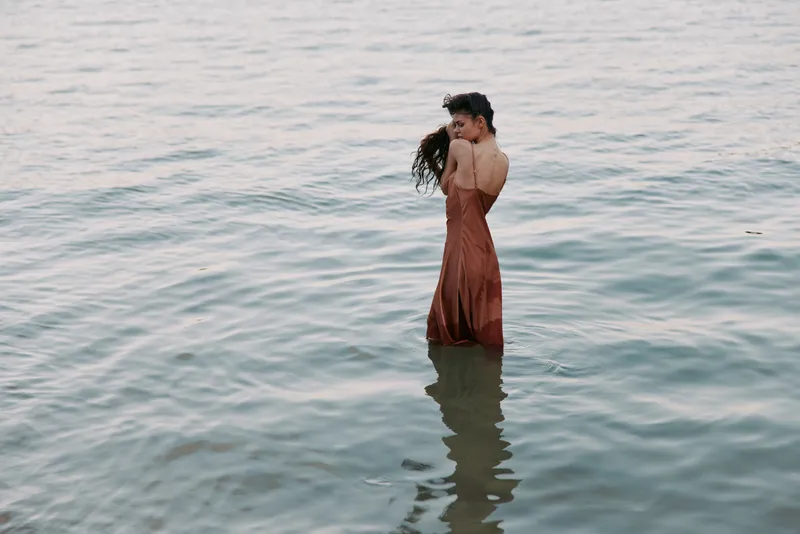 A solitary figure stands waist deep in calm water, wearing a flowing dress, captured in soft light with gentle ripples surrounding and a contemplative mood.