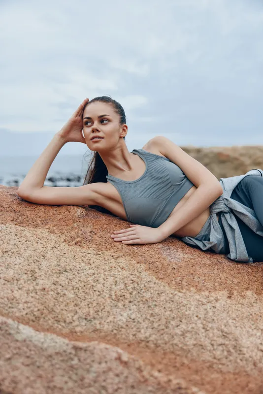 Fit young woman resting on a rock in activewear, showcasing a serene expression against a natural backdrop, emphasizing health and fitness in a coastal setting