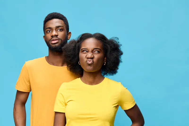 A young African couple in bright yellow t shirts, expressing playful emotions against a vibrant blue background, showcasing joy and spontaneity