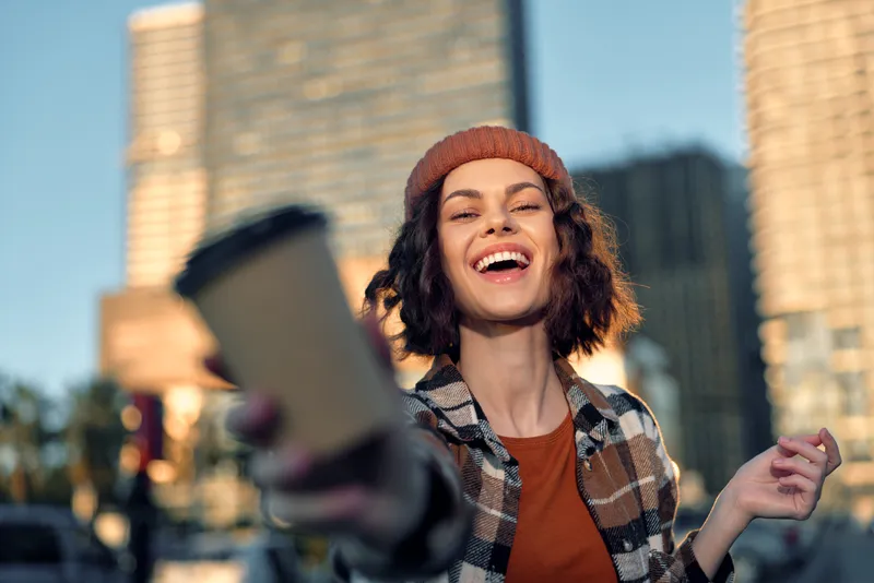 Woman smiling with coffee in city street at golden hour, candid outdoor portrait in beanie and plaid shirt, authenticity and emotional storytelling highlight mindful living lifestyle.