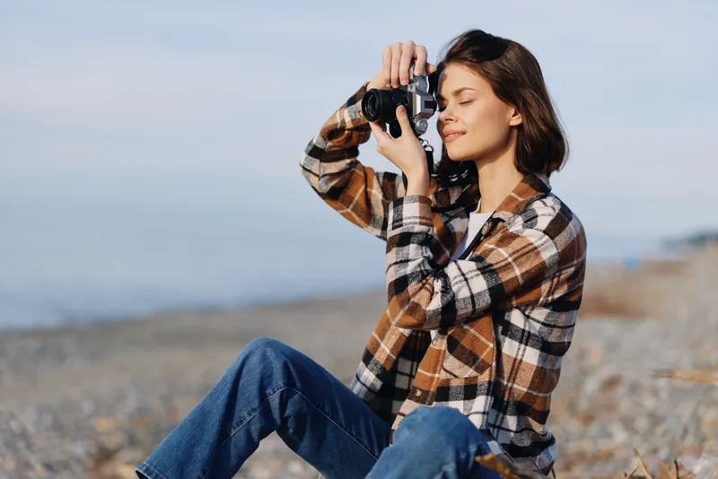 Photographer woman outdoors on a beach, capturing landscape with camera, wearing plaid shirt and jeans, focused on shot under natural light by the sea