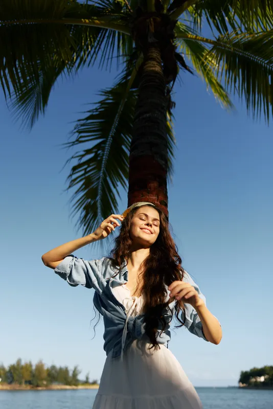Joyful woman stands under a tall palm, sunlit smile, breeze in hair, wearing a light summer dress. Tropical backdrop with clear blue sky and gentle water.
