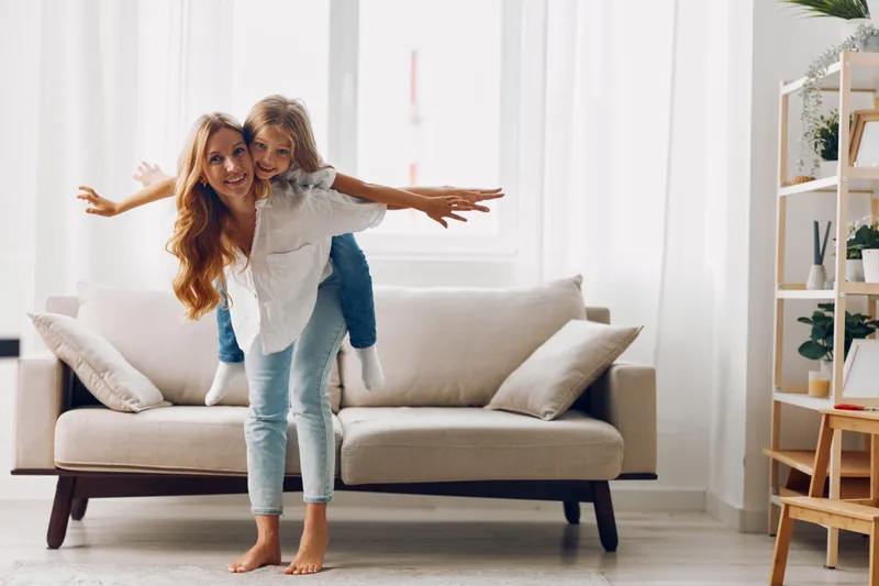 Мать и дочь радостно стоят на диване в гостиной, подняв руки вверх, празднуя вместе веселый момент
