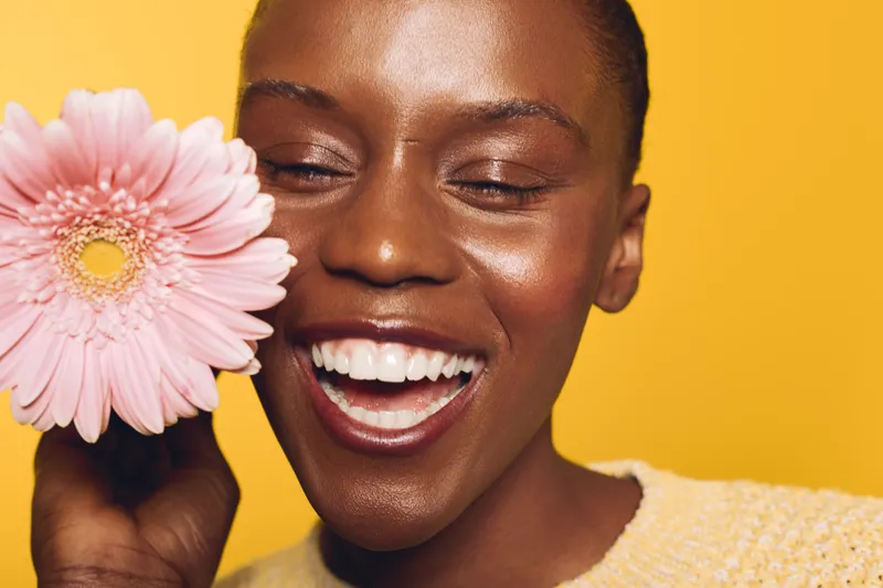 Happy African woman with dark skin laughs holding pink flower near face on yellow background. Concept of joy, natural beauty, skincare, and positive emotions in studio portrait.
