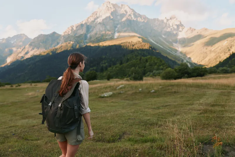 Young woman hiker with backpack walks on grassy field surrounded by mountains under blue sky, enjoying outdoor lifestyle and nature adventure in summer.