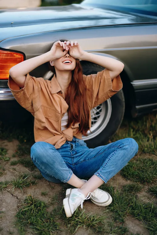 Young woman with long red hair laughing joyfully while sitting beside a vintage car, dressed casually in a brown shirt and blue jeans.