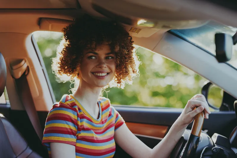 Young woman with curly hair sits behind the wheel of a car, smiling brightly as she drives, wearing a colorful striped shirt, with warm natural light streaming through the windows.