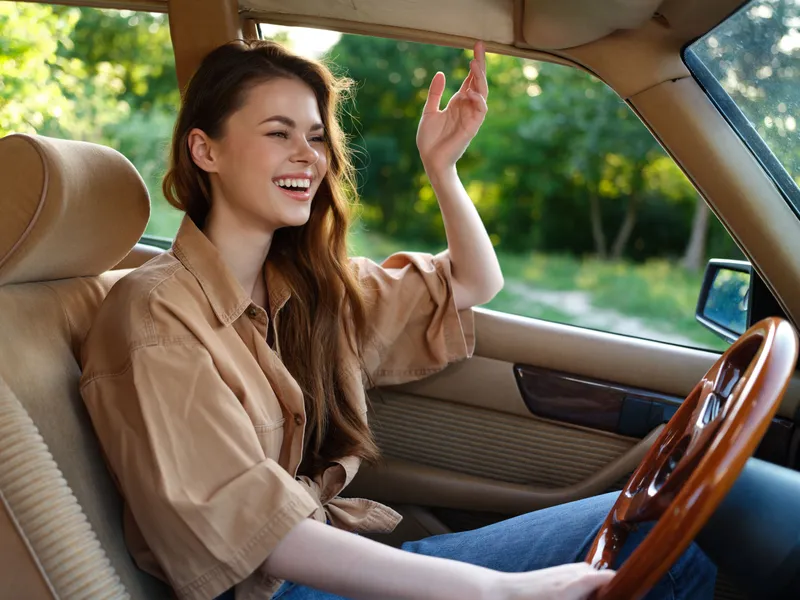 Joyful young woman driving in a vintage car, wearing casual attire, with a bright smile in a lush green background.
