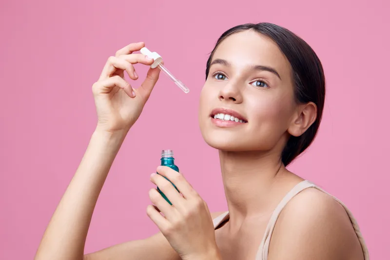 Young female applying serum, showcasing healthy skin and natural beauty against a pink background, emphasizing skincare and self care routine