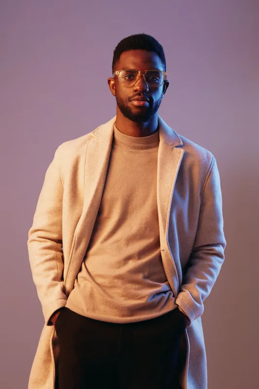 Stylish African man in a long coat and glasses, showcasing modern fashion against a vibrant purple background, embodying confidence and elegance