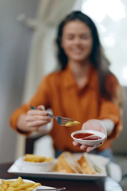 Счастливая женщина наслаждается едой, держа картофель фри с кетчупом, в оранжевом топе, в теплой и гостеприимной атмосфере, что подчеркивает привычки здорового питания
