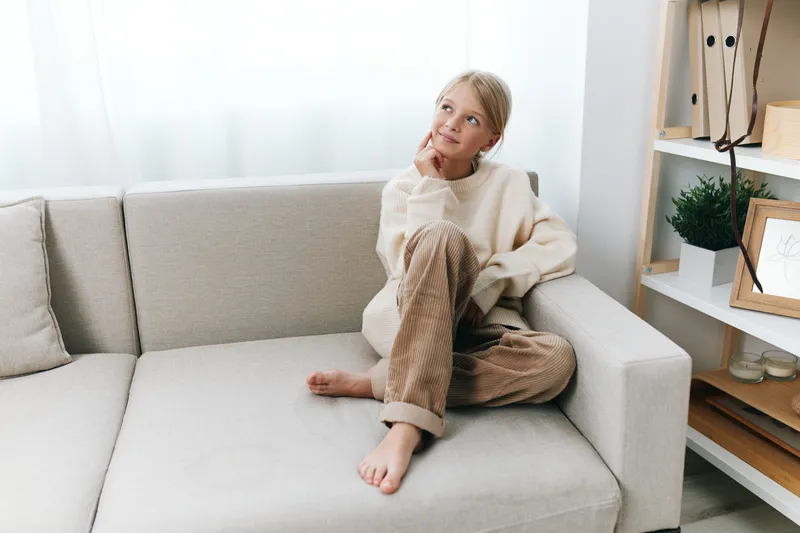 Bright indoor scene features a relaxed woman sitting on a pale sofa, wearing a cream sweater and striped pants, basking in soft natural light and a tranquil, cozy ambiance.