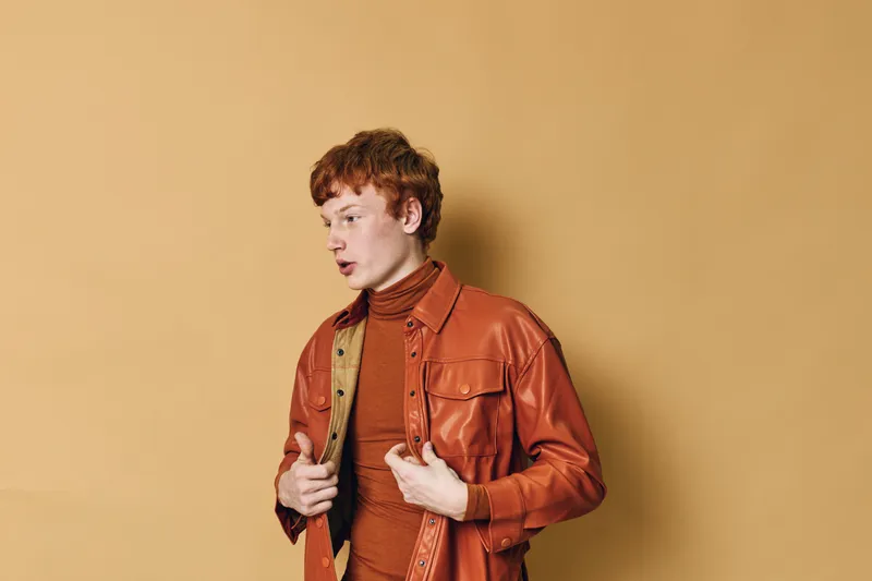 Young man with red hair wearing orange jacket and turtleneck poses against beige background looking sideways. Fashion, casual outfit, model, studio portrait and autumn style concept.