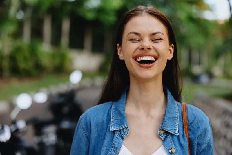Портрет брюнетки, которая улыбается, обнажая зубы, и идет на улице на фоне пальм в тропиках