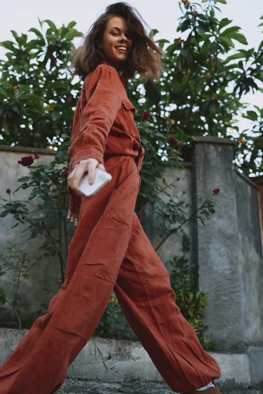 Elegant young woman in a rust jumpsuit strides toward the camera in a sunlit urban street, projecting confidence and a carefree, candid energy with a light breeze ruffling her hair