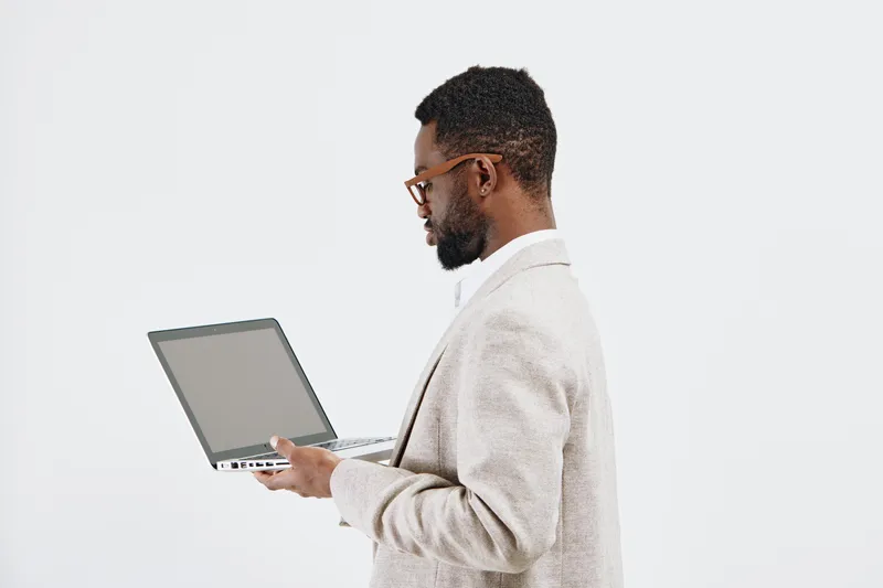 Thoughtful man in glasses and light sweater holding a laptop focusing on screen, isolated on plain white background, studio portrait. Technology, business, workspace concept