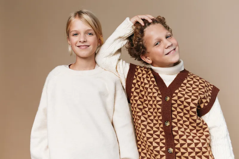 Two smiling children pose in cozy knitwear against a warm beige backdrop, showcasing soft textures and a relaxed, inviting fashion vibe for family scenes.